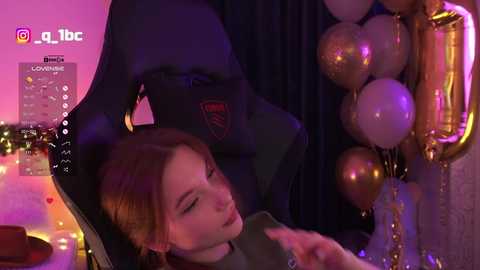 Video of a young woman with fair skin, red hair, and closed eyes, resting on a black gaming chair. The background features festive decorations, including gold balloons and strings of fairy lights.
