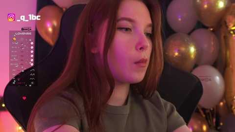 Video of a young woman with long brown hair, wearing a green shirt, sitting in a black gaming chair, surrounded by gold and white balloons, with purple and pink lighting.