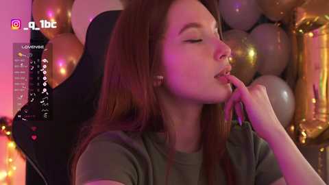 Video of a fair-skinned woman with long auburn hair, wearing a green t-shirt, touching her lips sensually, in a dimly lit room with gold balloons and purple lighting, looking relaxed and intimate.