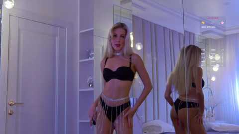 Video of a slim, blonde woman in black lingerie and sheer skirt standing confidently in a modern, minimalist bedroom with mirrored walls and soft lighting.