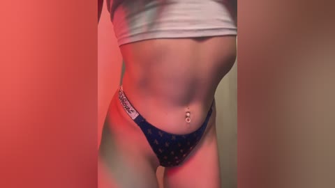 Media: Video of a light-skinned woman's torso, wearing a cropped white top and blue patterned panties, with a navel piercing, bathed in soft, warm lighting.