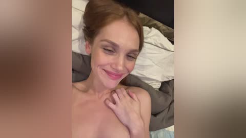 A video of a topless woman with fair skin, light brown hair, and closed eyes, lying on a bed with grey and white sheets, smiling.