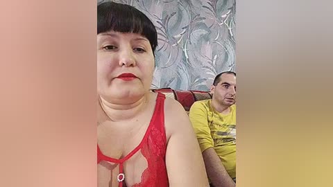 Video of a plus-sized woman with dark hair and a nose ring, wearing a red lace bra, sitting next to a man in a yellow shirt on a red couch.