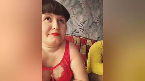 A video of a plus-size woman with pale skin, dark hair, and red lipstick, wearing a red lace bra, sitting on a couch with a patterned cushion, in a room with floral wallpaper.