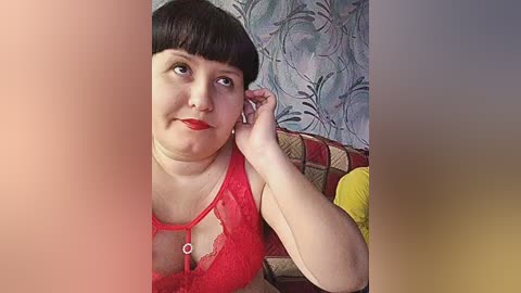 A video of a plus-sized woman with pale skin, dark hair, and red lipstick, wearing a red lace bra, sitting against a floral-patterned wall, adjusting a headphone.