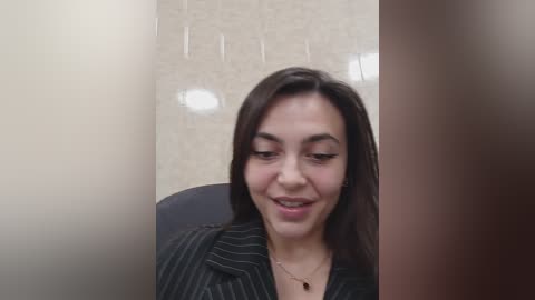 A video of a smiling young woman with dark hair and light skin, wearing a black pinstriped shirt, sitting in a beige-tiled bathroom.
