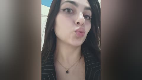 A close-up video of a young woman with fair skin, long dark hair, and a black pinstriped jacket, puckering her lips in a playful pose.