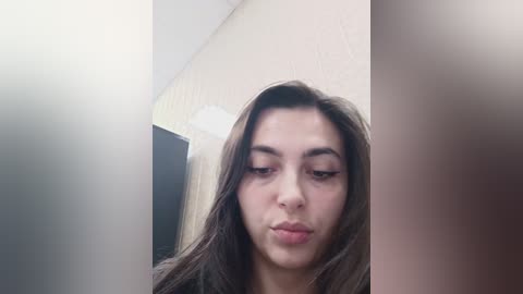 Video of a young woman with light brown skin, straight dark brown hair, and full lips, making a pouty face in a dimly lit room with beige walls.