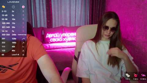 Video of a young woman with long brown hair and sunglasses, wearing a white t-shirt, sitting in a modern, pink-lit room with a neon sign behind her.