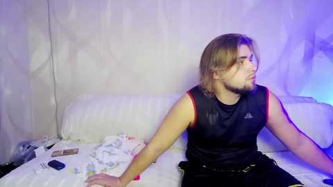 Media: Video of a young man with shoulder-length brown hair, wearing a black sports tank top, sitting on a bed with a white sheet. He has a serious expression. Background features a white wall and a blue light.