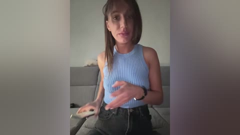 A video of a young woman with straight, light brown hair, wearing a light blue ribbed tank top and high-waisted jeans, seated on a grey couch, making a gesture with her right hand.