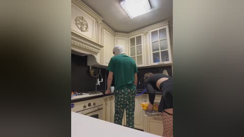 A video of an elderly man in green pajamas and a white cap, cooking in a beige kitchen with a modern design and white cabinets, while a younger woman in a black top and patterned pants stands by the counter.