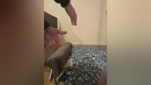 Media: Video of a person's hand pointing at a person lying on a bed with a green and white patterned blanket. The room has beige wallpaper and a wooden floor.