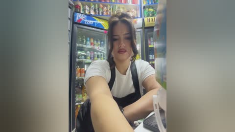 Media: Video of an Asian woman with dark hair in a white shirt and black apron, working in a well-lit convenience store. She has a serious expression and is reaching into a glass-fronted cooler.