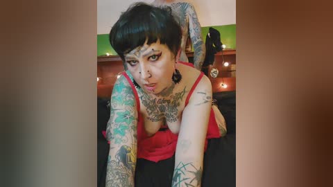Video of a tattooed woman with short black hair, wearing a red top, lying on a bed with a green wall behind her.