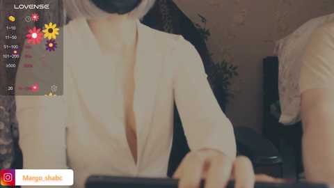 Video of a woman in a low-cut white blouse, with a black mask covering her face, sitting at a desk. Background shows a dimly lit room with a potted plant and a black object.