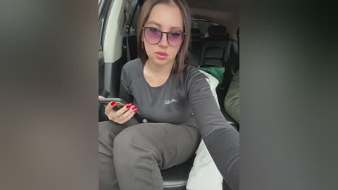Media: Video of a woman with fair skin, dark hair, wearing purple sunglasses, black shirt, and gray pants, sitting in a car, holding a phone, with blurred background.