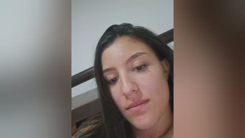 Video of a young woman with light skin, long black hair, and brown eyes, looking slightly tired. She is indoors, leaning against a wooden headboard.