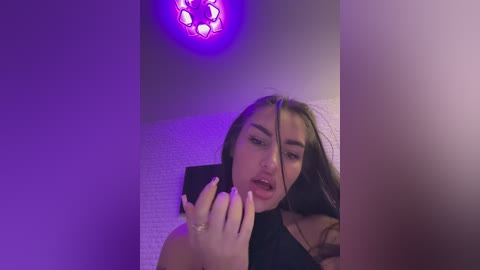Media: A video of a young woman with long brown hair, wearing a black top, taking a selfie with a smartphone. The background features a purple-lit chandelier and textured white wall.