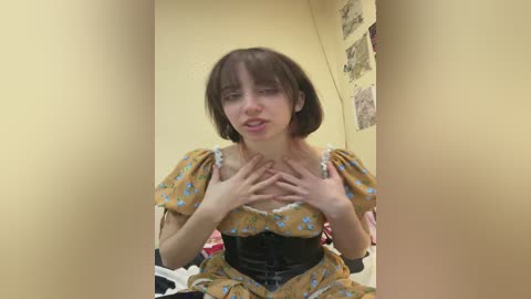Media: A video of a young woman with pale skin and short brown hair, dressed in a yellow floral dress, touching her chest with her hands, in a bedroom with a beige wall and a poster.