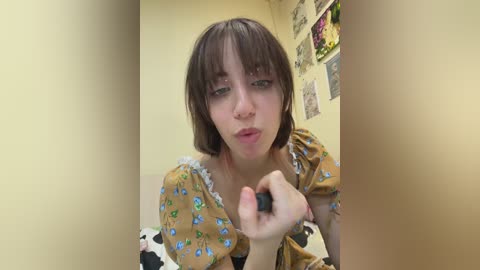 Media: Video of a young woman with short brown hair and a cow-print dress, making a silly face with her hand. Background features a beige wall with various framed pictures and a floral arrangement.