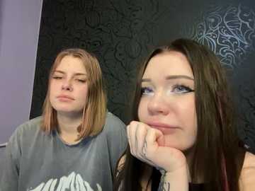 Media: Video of two young women with straight, shoulder-length hair, one in a grey T-shirt, the other in a black tank top, both with tattoos on their forearms, leaning against a dark, ornate wall.