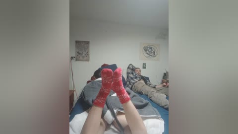 Video of a dimly lit bedroom with a person's feet in red and white patterned socks on a bed. A man in a striped shirt and pants is sitting on another bed.