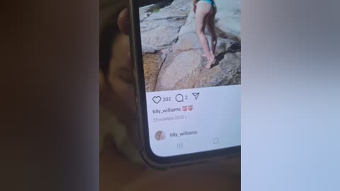 Media: Video of a smartphone screen displaying a profile photo of a woman in a bikini, sitting on a rock, with a dimly lit, blurry background.