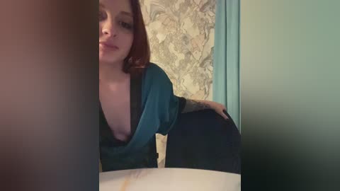 Media: Video of a curvy, tattooed woman with shoulder-length red hair, wearing a teal blouse, leaning over a marble table, reflected in a mirror with a floral-patterned wall in the background.