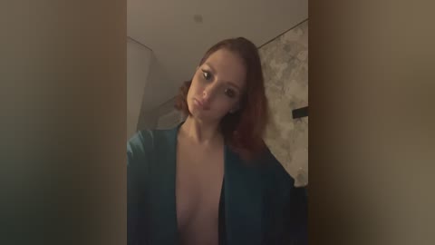 Media: A video of a fair-skinned woman with shoulder-length red hair, wearing a teal blouse, revealing cleavage, standing in a dimly lit, modern bathroom with marble tiles and a mirror.