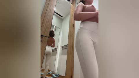 Media: A video of a woman standing in a dressing room, facing a full-length mirror, wearing white high-waisted leggings, her breasts exposed, her arms crossed.