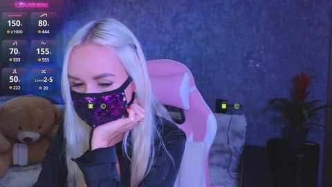 Video of a blonde woman in a black face mask, pink gaming chair, and plush toy, in a dimly lit room with gaming overlays on screen.