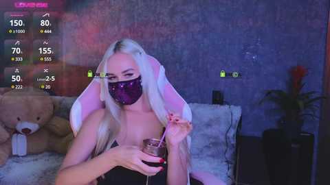 Video of a blonde woman in a dark room, wearing a black face mask, holding a glass of wine, with a teddy bear and a potted plant in the background.