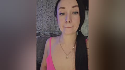 Video of a young Caucasian woman with fair skin, black hair, wearing a pink spaghetti-strap top and a gold necklace, smiling against a textured grey wall background.