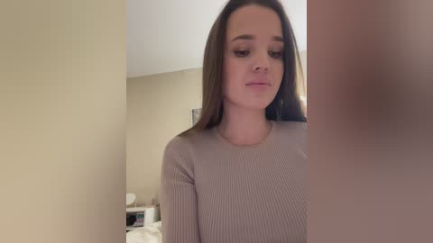 Media: Video of a young Caucasian woman with long brown hair, wearing a ribbed gray sweater, standing in a dimly lit room with beige walls.