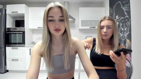 Media: Video of two young women in a modern kitchen: a blonde with long hair in a white crop top, and a brunette in a black crop top, both looking at a smartphone.