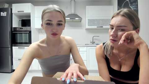 Media: Video of two women in a modern kitchen: a slender blonde in a strapless top, and a brunette in a black tank top, both looking annoyed.