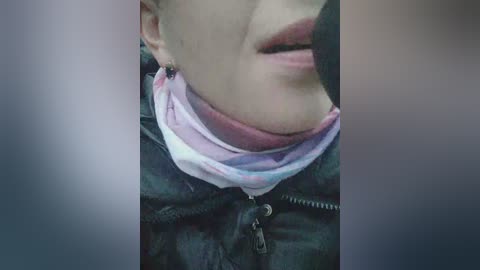 Video of a person with light skin, wearing a pink and purple face mask, a dark jacket, and visible nose stud. The background is blurred.