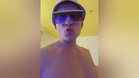 Media: Video of a shirtless, light-skinned man with blue-tinted 3D glasses, kissing the air. He wears a light gray beanie. Background features a yellow ceiling and white walls.