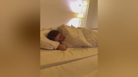 Media: A video of a woman with brown hair, sleeping under white sheets, in a dimly lit bedroom with a nightstand lamp.