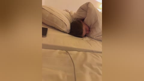 Media: Video of a man lying on a bed with beige sheets, head covered with a pillow, wearing a white shirt, holding a smartphone.