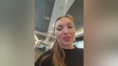 A candid video of a blonde woman with fair skin and pink lips, wearing a black top, in a modern, brightly lit interior with tiled ceiling and blurred background.