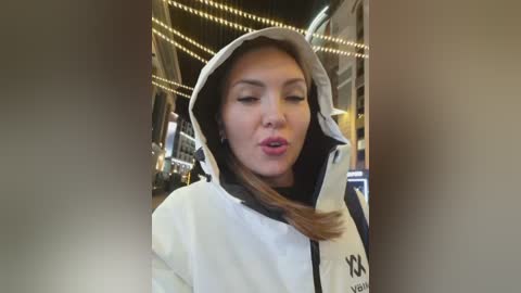 A video of a Caucasian woman with light skin and straight brown hair, wearing a white and black hoodie, blowing a kiss while indoors, surrounded by warm, ambient lighting.