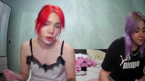 Media: Video of two young women with vibrant red and purple hair, one in a black camisole, the other in a black Puma t-shirt, in a messy bedroom with light blue wallpaper.