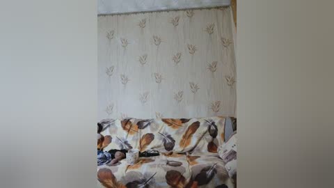 A video of a cozy, cluttered room with a white sofa covered in a floral pattern featuring brown, beige, and gray leaves. A sheer curtain with a similar leaf design hangs behind, and a TV remote rests on the sofa.
