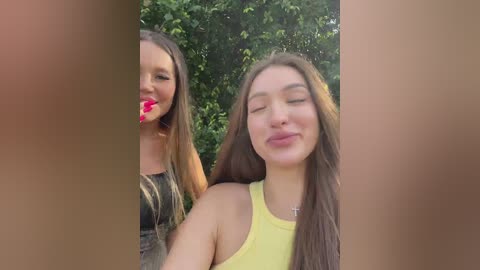 Media: Video of two young women with fair skin, long brown hair, and medium builds. One is wearing a black top, the other a yellow tank top. They are smiling and playfully sticking their tongues out. Green foliage in the background.