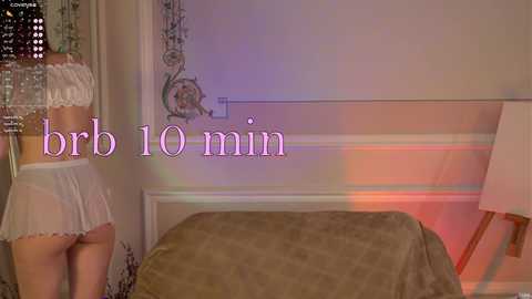 A video of a woman in white lingerie and a short skirt, standing beside a bed with a beige blanket, in a dimly lit room with a glowing lamp.