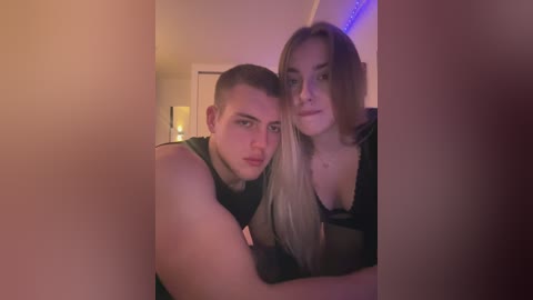 Media: Video of a young couple in a dimly lit room; the man with short hair, wearing a tank top, and the woman with long blonde hair, wearing a black bra, both appear casual and relaxed.