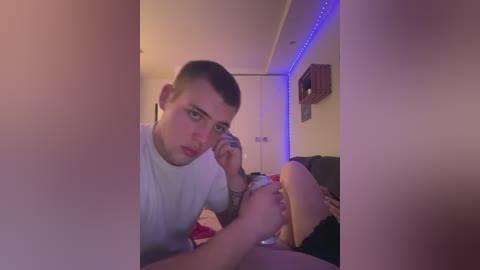 Media: A young, pale-skinned man with short brown hair and a white t-shirt leans over a nude woman with fair skin, applying nail polish to her toes in a dimly lit room with blue LED lights.