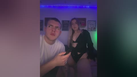 Media: Video of a young man with short hair in a white T-shirt, looking shocked, and a blonde woman in black lingerie, both indoors with purple lighting.